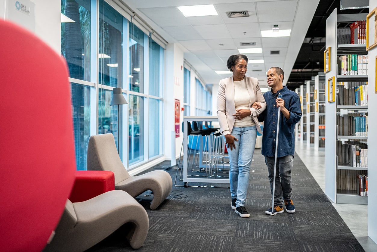 Teacher walks through the university library with visually impaired student, using a walking cane. Teaching students with special needs. 